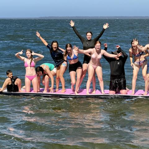 Group of campers standing on a giant stand up paddleboard, smiling at the camera as they float on the water