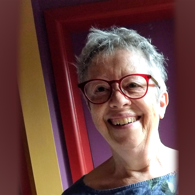 Person smiling at the camera with ruffled grey hair and red rimmed glasses